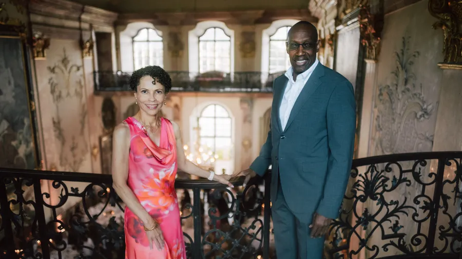 two nicely-dressed individuals stand on a balcony overlooking a grandiose hall