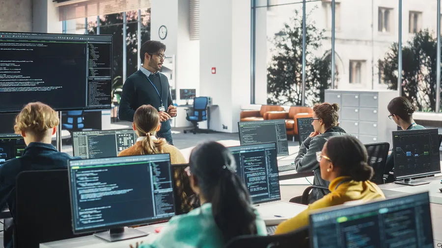 A teacher stands in front of a room of students who are seated at computers