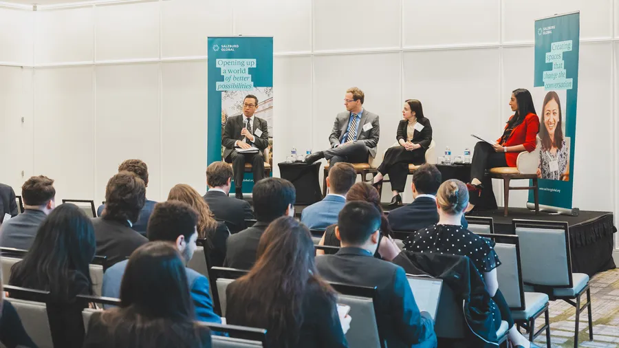An image of four individuals seated on a stage in front of an audience. A man on the left speaks into a microphone.