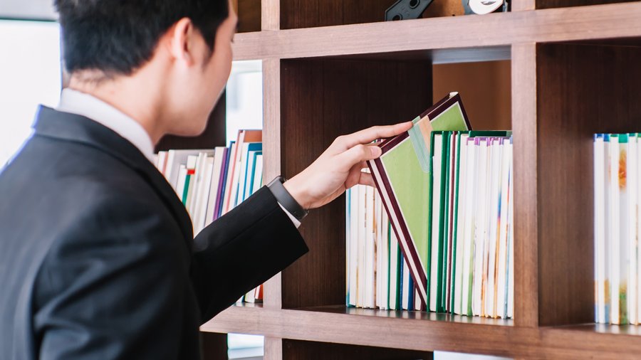 A man wearing with dark hair wearing a suit reachs to remove a book from a bookshelf.