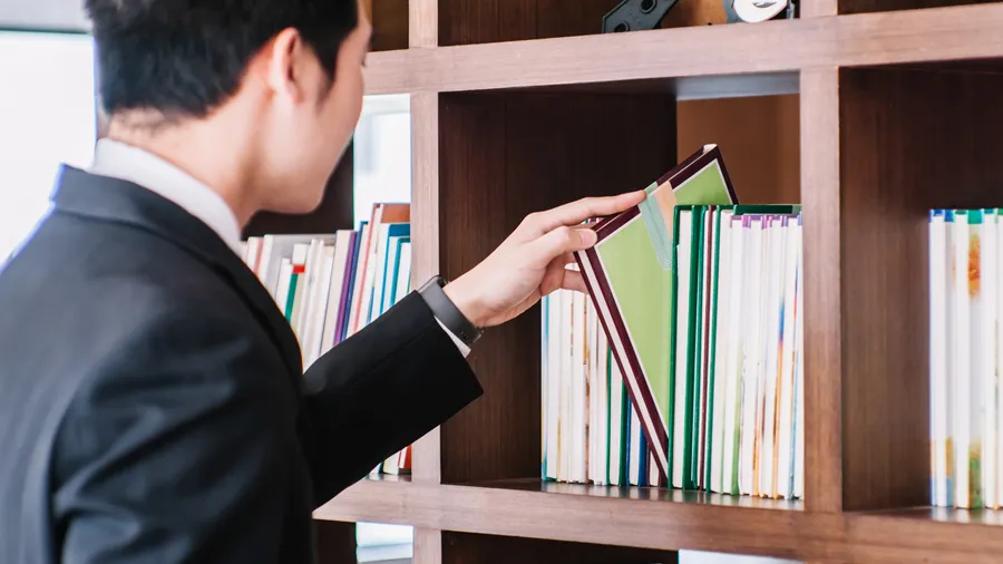 A man wearing with dark hair wearing a suit reachs to remove a book from a bookshelf.