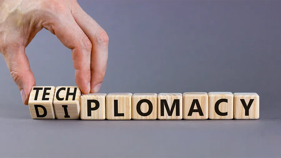 The image depicts a hand adjusting or placing wooden blocks. These blocks spell out the word "DIPLOMACY" but are partially modified on the left side to emphasize the word "TECH" within it. The background is a simple gray, and the focus is on the juxtaposition of the terms "tech" and "diplomacy," suggesting the concept of "tech diplomacy" or the intersection of technology and diplomatic relations.