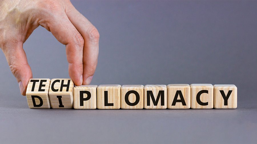 The image depicts a hand adjusting or placing wooden blocks. These blocks spell out the word "DIPLOMACY" but are partially modified on the left side to emphasize the word "TECH" within it. The background is a simple gray, and the focus is on the juxtaposition of the terms "tech" and "diplomacy," suggesting the concept of "tech diplomacy" or the intersection of technology and diplomatic relations.