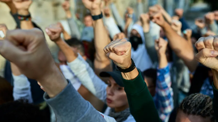 An eye-level shot captures a large gathering of people, their fists raised high in what appears to be a collective show of solidarity or protest. The crowd members are wearing a variety of casual clothing, including t-shirts, jackets, and caps. The background suggests an urban setting, with blurred buildings in the distance.