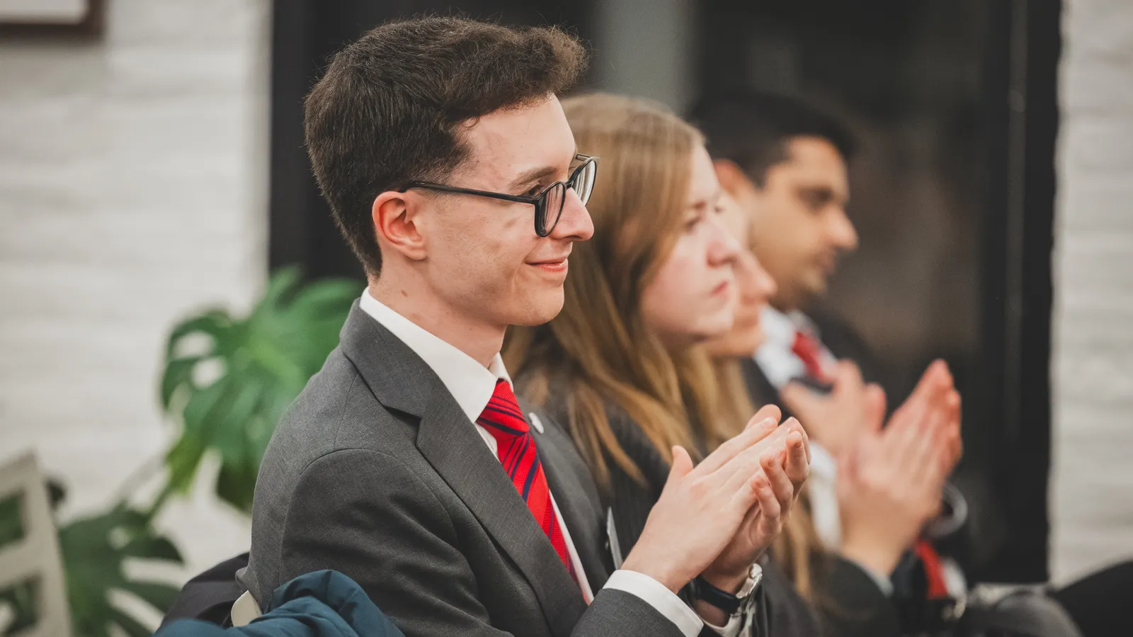 a man wearing a suit and glasses claps while looking off-camera to the right