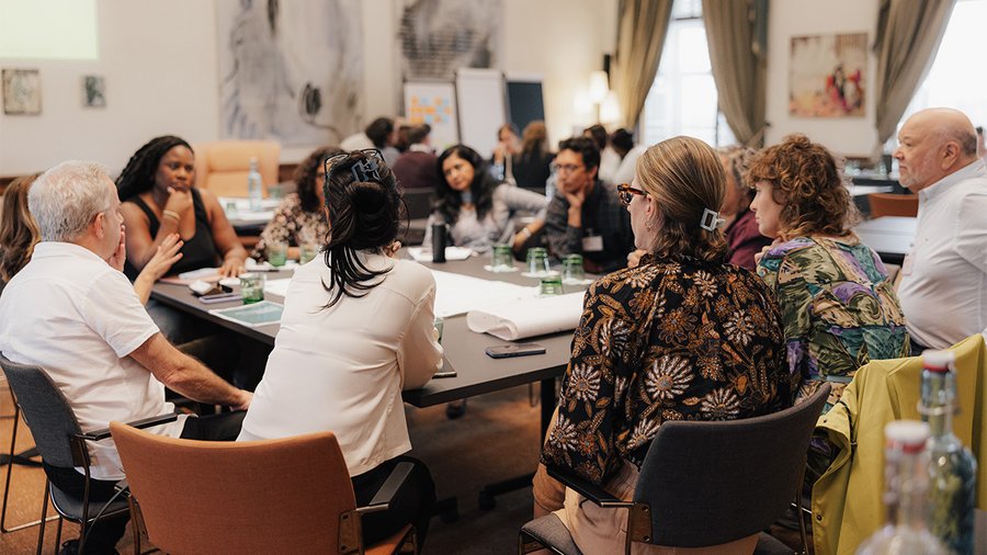 A group of people sits around a table in deep discussion, with the backs of several women to the viewer. 
