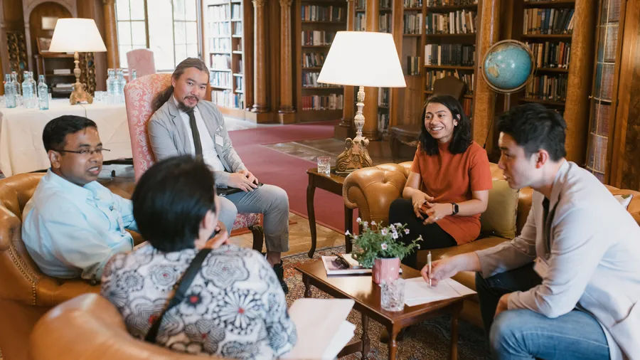 A diverse group of individuals engaged in a collaborative discussion, seated in a cozy library setting with wooden bookshelves, a globe, and warm lighting creating an inviting atmosphere.