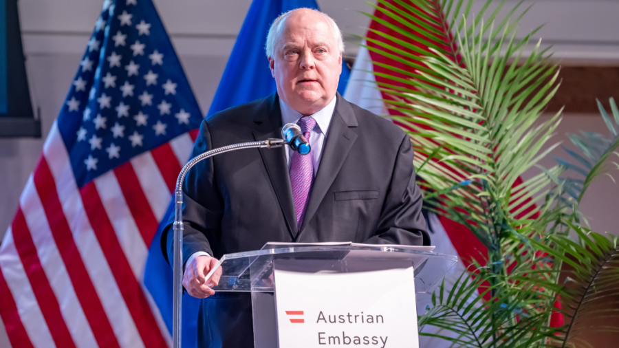 Hugh Verrier, a man with white hair, stands at a podium delivering a lecture in the Austrian Embassy in Washington. In the background are American and Austrian flags. 